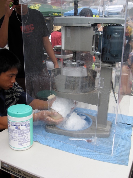 A young man shaves a block of ice using a heavy-duty machine and molds a mound of the shavings into a small plastic cup.