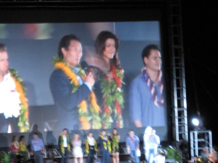 Daniel Dae Kim, wearing a golden-yellow lei and ti leaves and flanked by costars Scott Caan and Grace Park, speaks onstage to the crowd